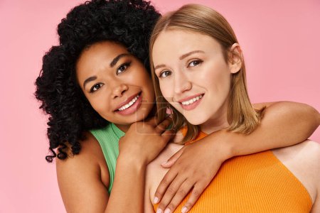 Two young women embrace warmly against a soft pink backdrop.