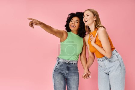 Two attractive diverse women pointing at something, standing next to each other.