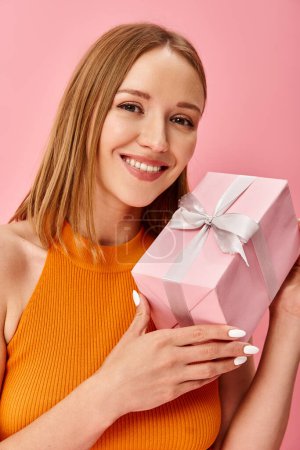 Une femme dans un haut orange tient joyeusement une boîte-cadeau rose.