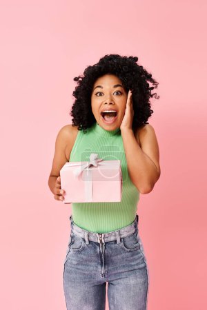 A diverse, attractive woman in casual attire, holding a pink gift box with a surprised look on her face.