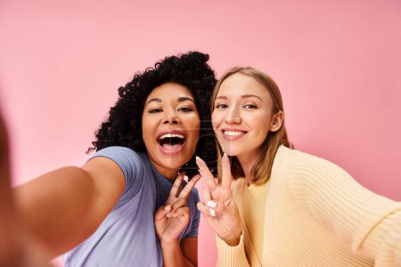 Two women in casual attire pose for a selfie in front of a pink wall.