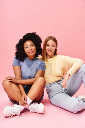 Zwei unterschiedliche Frauen in kuscheligen Outfits sitzen auf dem Boden und posieren für ein Foto.
