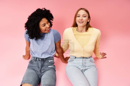Two diverse women in cozy attire sitting gracefully on a pink background.