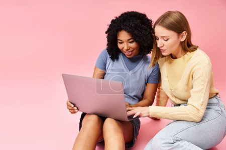 Foto de Two diverse women in casual attire are sitting on the ground, focused on a laptop. - Imagen libre de derechos