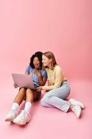 Photo for Two diverse women in casual attire sit on the floor, engrossed in using a laptop. - Royalty Free Image