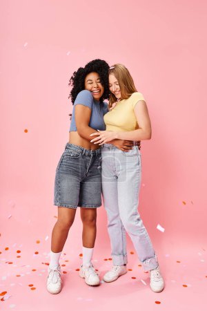 Two attractive diverse women embrace warmly in front of a soft pink backdrop.