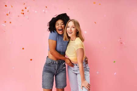 Zwei attraktive junge Frauen in kuscheliger Kleidung stehen nebeneinander vor einem leuchtend rosa Hintergrund.