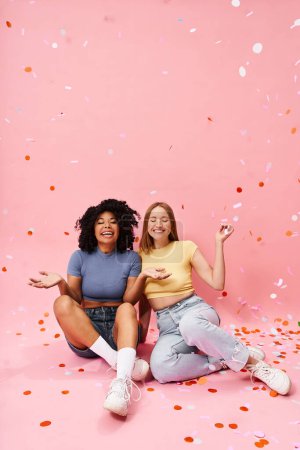 Two diverse women in cozy casual attire sitting on the ground surrounded by confetti.