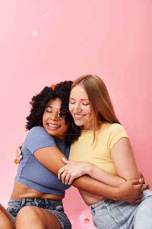 Two attractive women hugging warmly on a soft pink background.