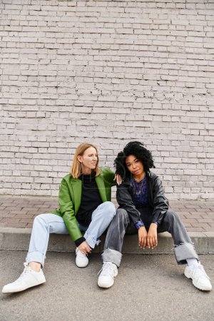 Two diverse women in casual attire sit on curb by brick wall.