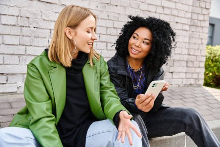 Photo for Two diverse women in casual attire sit on a bench, engrossed in a cell phone. - Royalty Free Image