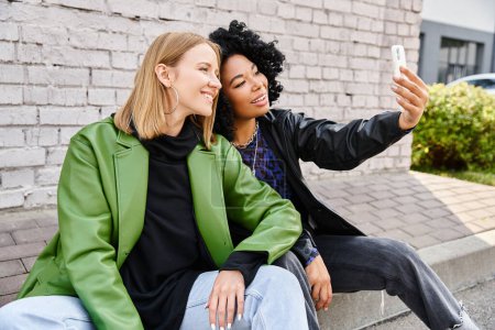 Two attractive diverse women in cozy casual attire sit closely together, enjoying each others company.