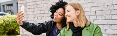 Téléchargez les photos : Deux jolies femmes diverses en tenue décontractée prenant un selfie avec un téléphone portable. - en image libre de droit