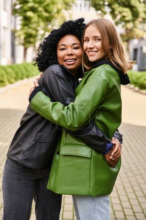 Two attractive diverse women warmly hugging on a sidewalk.