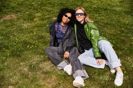 Foto de Dos mujeres diversas en traje casual relajarse en un campo verde vibrante. - Imagen libre de derechos