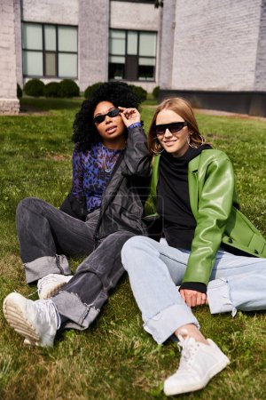 Dos mujeres diversas en traje casual se sientan en la hierba frente a un gran edificio.