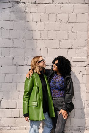 Two attractive diverse women in cozy casual attire standing near a brick wall.