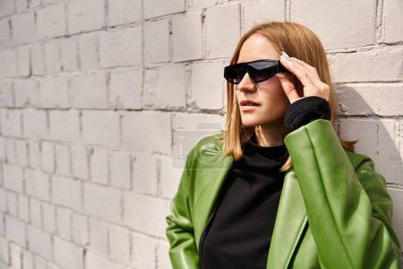 Stylish woman in green jacket and sunglasses leaning against a brick wall.
