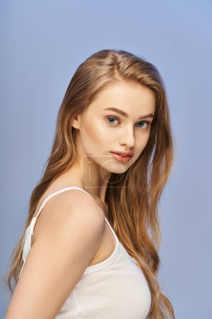 A young, blonde woman with long hair wearing a white tank top poses elegantly in a studio setting.