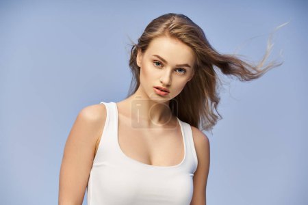 A young, blonde woman exudes elegance as she poses for a picture in a studio setting while wearing a white tank top.