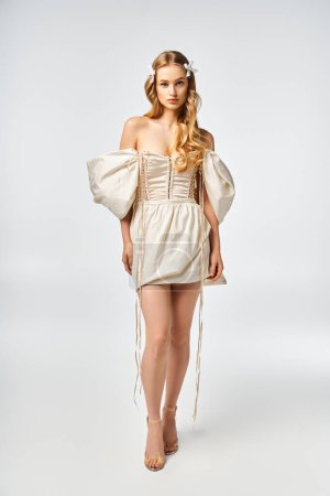 Eine junge blonde Frau steht elegant in einem weißen Kleid in einem Studio-Ambiente und strahlt Anmut und Charme aus.