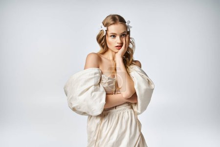 Eine junge blonde Frau strahlt Eleganz in einem Studio-Setting aus, trägt ein weißes Kleid und posiert für ein Foto.