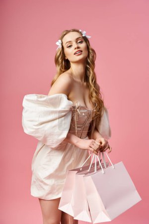 Foto de Joven, mujer rubia en un vestido blanco con gracia sostiene coloridas bolsas de compras en un entorno de estudio. - Imagen libre de derechos