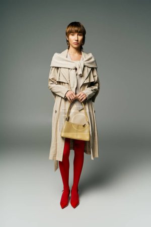 A stylish young woman with short hair poses in a trench coat and bright red tights in a studio setting.