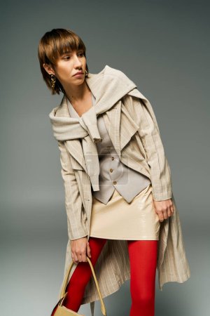 A chic young woman with short hair, donning tights and a coat, holds a fashionable handbag in a studio setting.