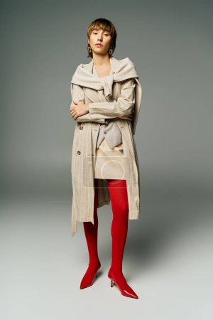 A young woman with short hair is strikingly dressed in a trench coat and vibrant red tights in a studio setting.