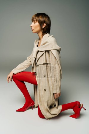 A young woman with short hair wearing a trench coat and red tights, exuding mystery and allure in a studio setting.