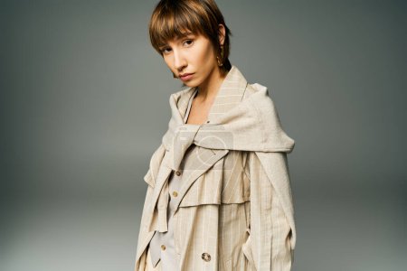 A stylish young woman with short hair poses confidently in a trench coat for a portrait in a studio setting.