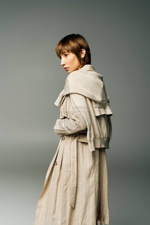 A young woman with short hair stands confidently in a trench coat against a solid gray background.