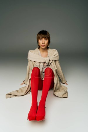 A young woman with short hair elegantly sits on the floor wearing striking red stockings in a studio setting.