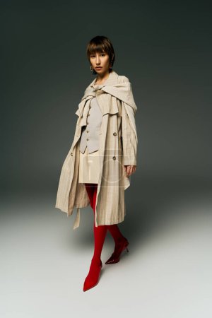 Eine stylische junge Frau im Trenchcoat präsentiert ihren einzigartigen Stil mit auffälligen roten Strumpfhosen im Studio-Ambiente.