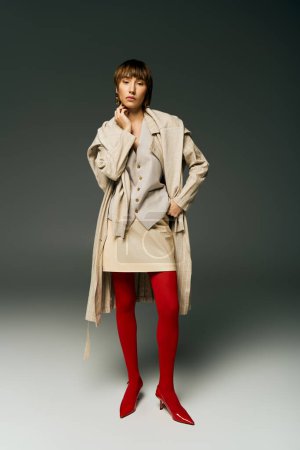 A stylish young woman in a trench coat and red tights strikes a pose in a studio setting, exuding confidence and sophistication.