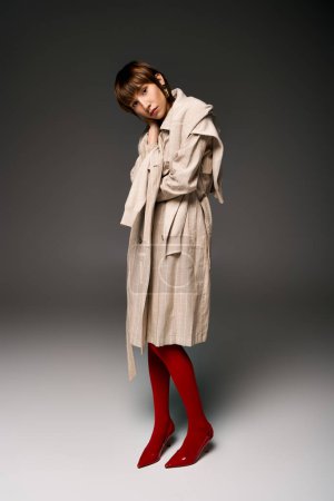 A young woman with short hair standing in a trench coat and red socks in a studio setting.