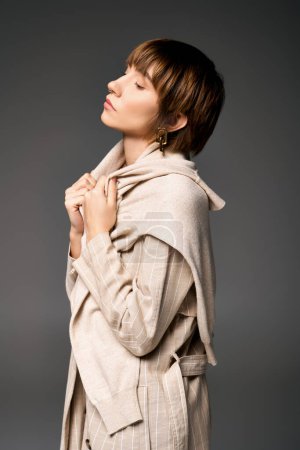 Téléchargez les photos : Une jeune femme aux cheveux courts pose gracieusement dans un manteau élégant, respirant la confiance dans un décor de studio. - en image libre de droit