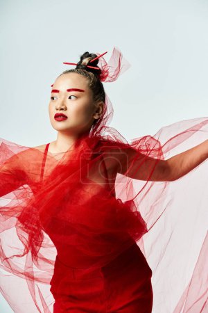 An attractive Asian woman in a vibrant red dress poses gracefully with a veil on her head.