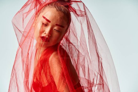 Femme asiatique intrigante dans une robe rouge frappante pose avec un voile sur la tête.