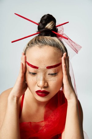 Une femme asiatique frappante ornée de maquillage rouge et de vêtements vibrants, tient dramatiquement sa tête.