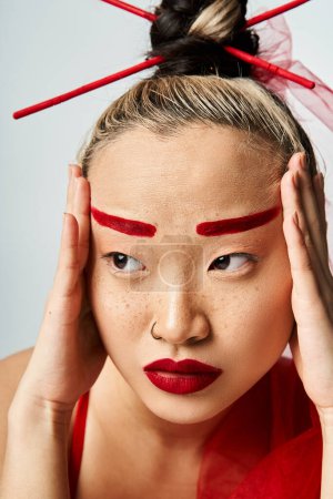 Téléchargez les photos : Une femme asiatique attrayante avec un maquillage rouge tenant sa tête de façon spectaculaire. - en image libre de droit