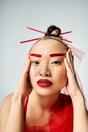 Asian woman in vivid attire, with red makeup, holds her head in a dramatic pose.