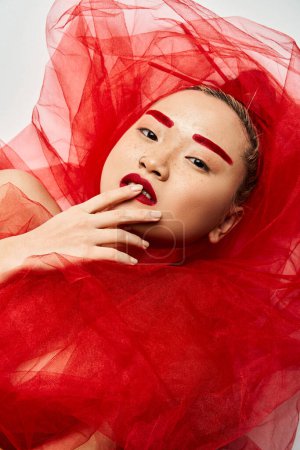 Foto de Impresionante mujer asiática en vibrante maquillaje rojo y vestido golpea una pose cautivadora. - Imagen libre de derechos