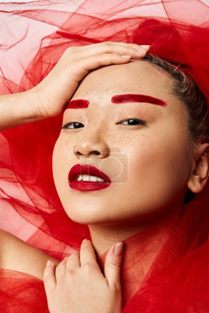 Une femme asiatique ornée de maquillage rouge et d'un voile rouge pose dynamiquement.