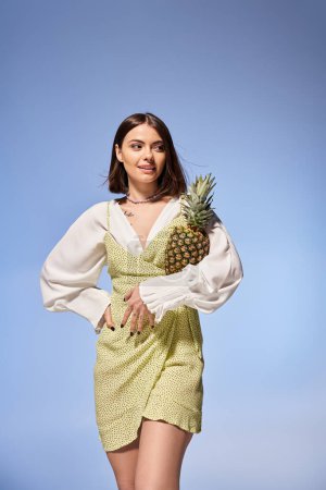 Photo for Beautiful young woman in a flowing dress gracefully holds a ripe pineapple in a vibrant studio setting. - Royalty Free Image