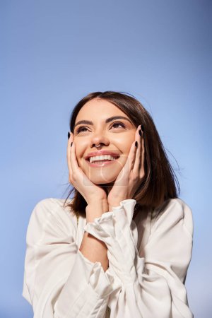 A young woman with brunette hair smiles joyfully, hands resting gently on her face in a studio setting.