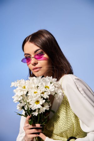 Téléchargez les photos : Une jeune femme aux cheveux bruns portant des lunettes de soleil tenant un bouquet de marguerites dans un cadre de studio serein. - en image libre de droit