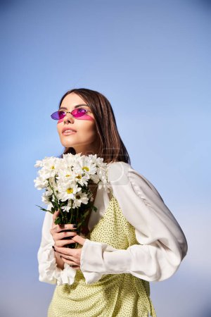 A young woman with brunette hair wearing sunglasses, holding a bouquet of daisies in a serene studio setting.