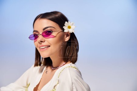 A stylish young woman with sunglasses and a flower in her hair strikes a pose in a studio setting.